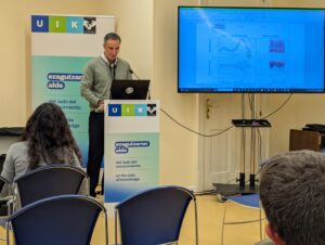 Guillermo Conde who si a representative from STT SYSTEMS, is giving a presentation, standing behind a podium with the "UIK" logo. He is speaking to a small audience. The presentation is likely about the company's 3DMA and ISEN movement analysis systems. A large screen to the right displays technical graphs and data related to movement ("Ángulo en el ciclo de marcha" - Angle in the gait cycle, and "Ángulo vs. Tiempo" - Angle vs. Time). A banner behind the presenter features the slogan "on the side of knowledge.