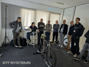 A female instructor sitting in front of a laptop gives an explanation to a group of men standing around a bicycle mounted on an indoor trainer.