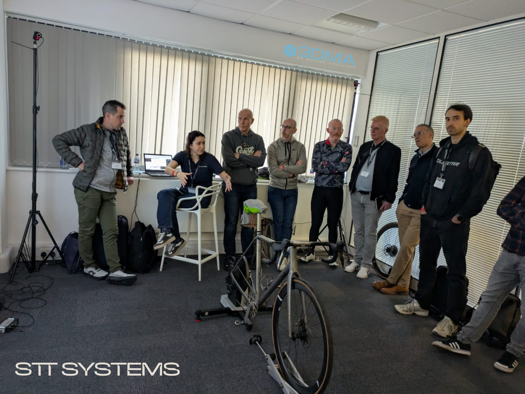 A female instructor sitting in front of a laptop gives an explanation to a group of men standing around a bicycle mounted on an indoor trainer.