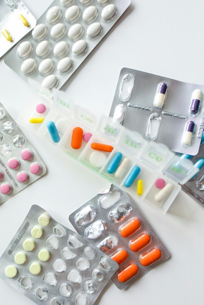 Pastillero semanal lleno de cápsulas de colores rodeado de varios blísteres de medicamentos sobre una mesa blanca.
