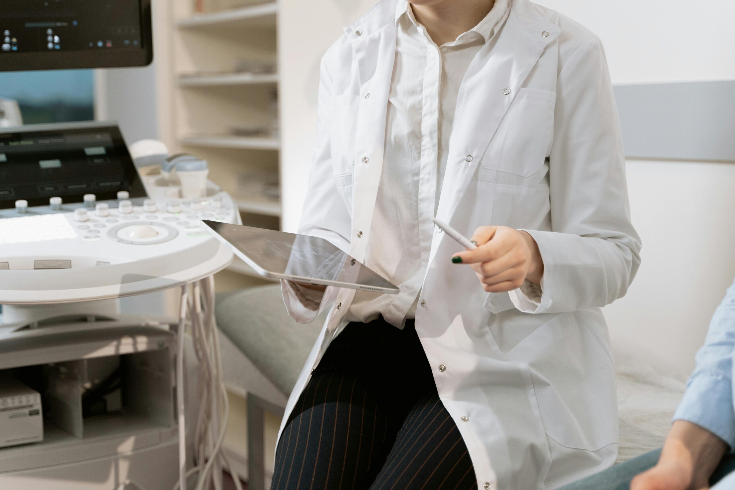Doctora con bata blanca sentada sosteniendo una tablet durante una consulta con un paciente.