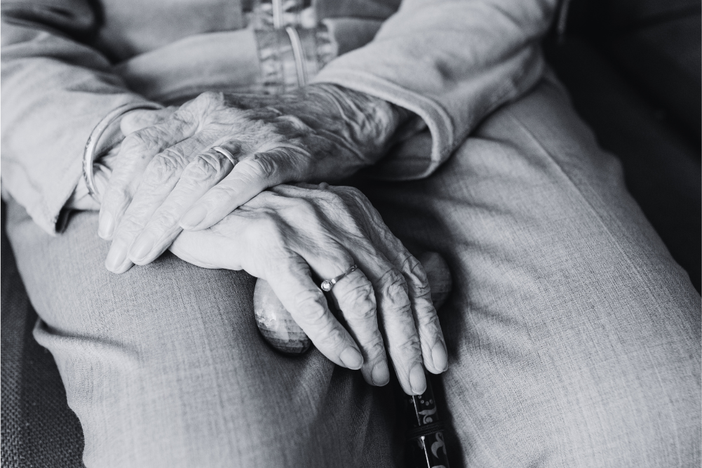 Foto en blanco y negro detallada y en primer plano de las manos profundamente arrugadas y envejecidas de una persona mayor, que llevan anillos y una pulsera sencilla, descansando sobre un bastón de madera en su regazo.