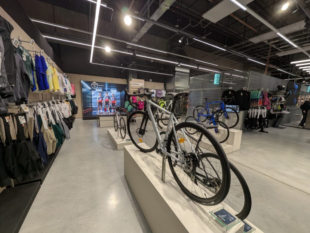 Pasillo de la tienda Van Rysel con bicicletas expuestas en pedestales y ropa ciclista colgada.