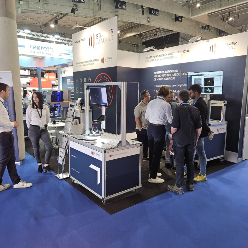 ALT (Texto Alternativo): Vista general del stand de STEMMER IMAGING en Advanced Factories, mostrando múltiples estaciones de demostración de visión artificial y un grupo numeroso de personas interactuando con el equipo y los representantes.