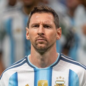 A close-up photograph of soccer player Lionel Messi. He has brown hair parted to the side, a full beard, and a serious expression looking straight ahead. He is wearing an Argentina national team jersey with black and light blue horizontal stripes. The white Adidas logo on the right side of his chest and the gold AFA (Argentine Football Association) crest with two stars on the left are clearly visible. The background is out of focus.