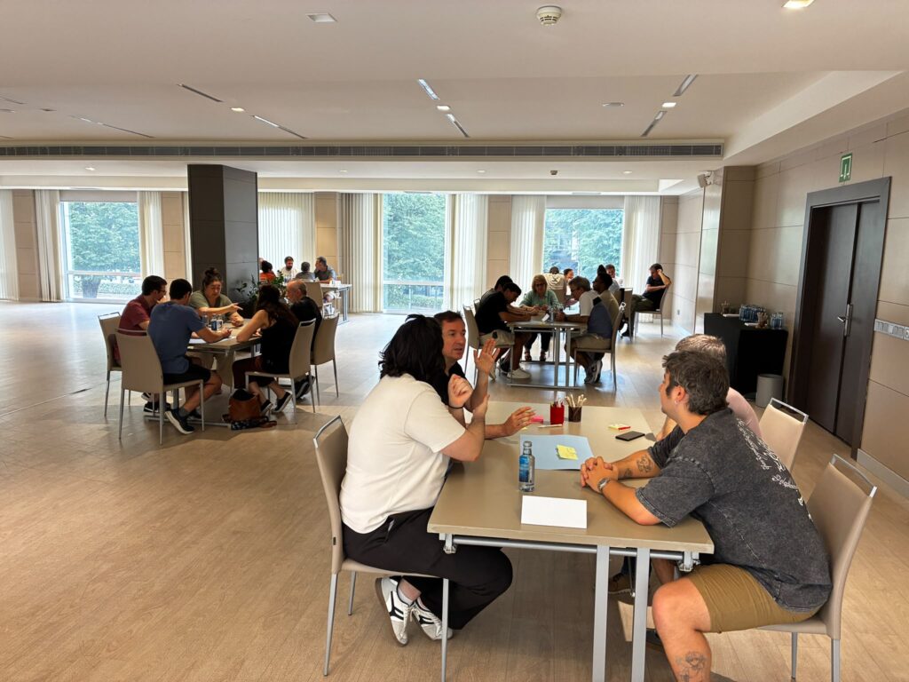 A wide shot of a bright meeting room where several small groups of people are sitting at tables, engaged in discussion during a collaborative workshop.