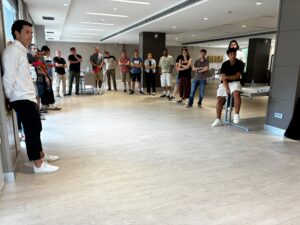 A group of casually dressed people gathered in a large room, standing in a wide circle and listening attentively during a meeting or workshop.