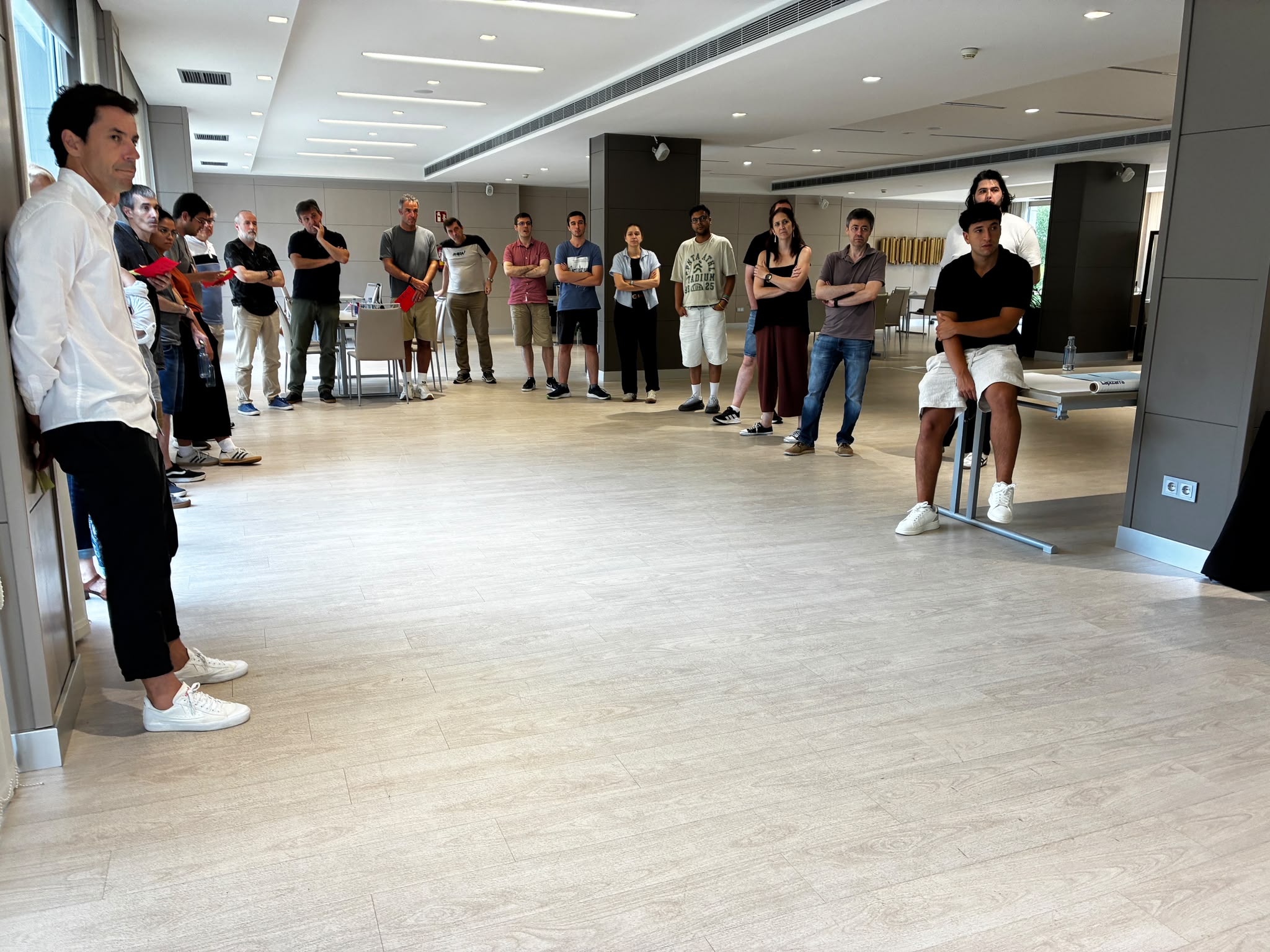 A group of casually dressed people gathered in a large room, standing in a wide circle and listening attentively during a meeting or workshop.