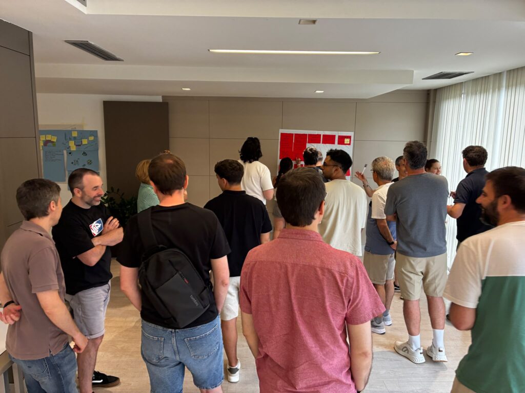 A group of people gathered around a wall, looking at a large red board with sticky notes during a collaborative brainstorming or workshop session.