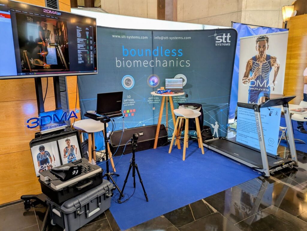 Stand de exposición de STT Systems en el que se muestra tecnología biomecánica, equipado con cámaras de captura de movimiento, una cinta de correr y monitores que muestran el software de análisis.